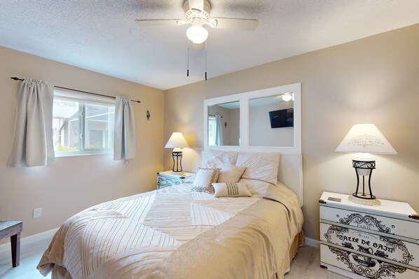 A bedroom with a bed, bedside tables, a ceiling fan, a window with curtains, and a dresser with decorative items.