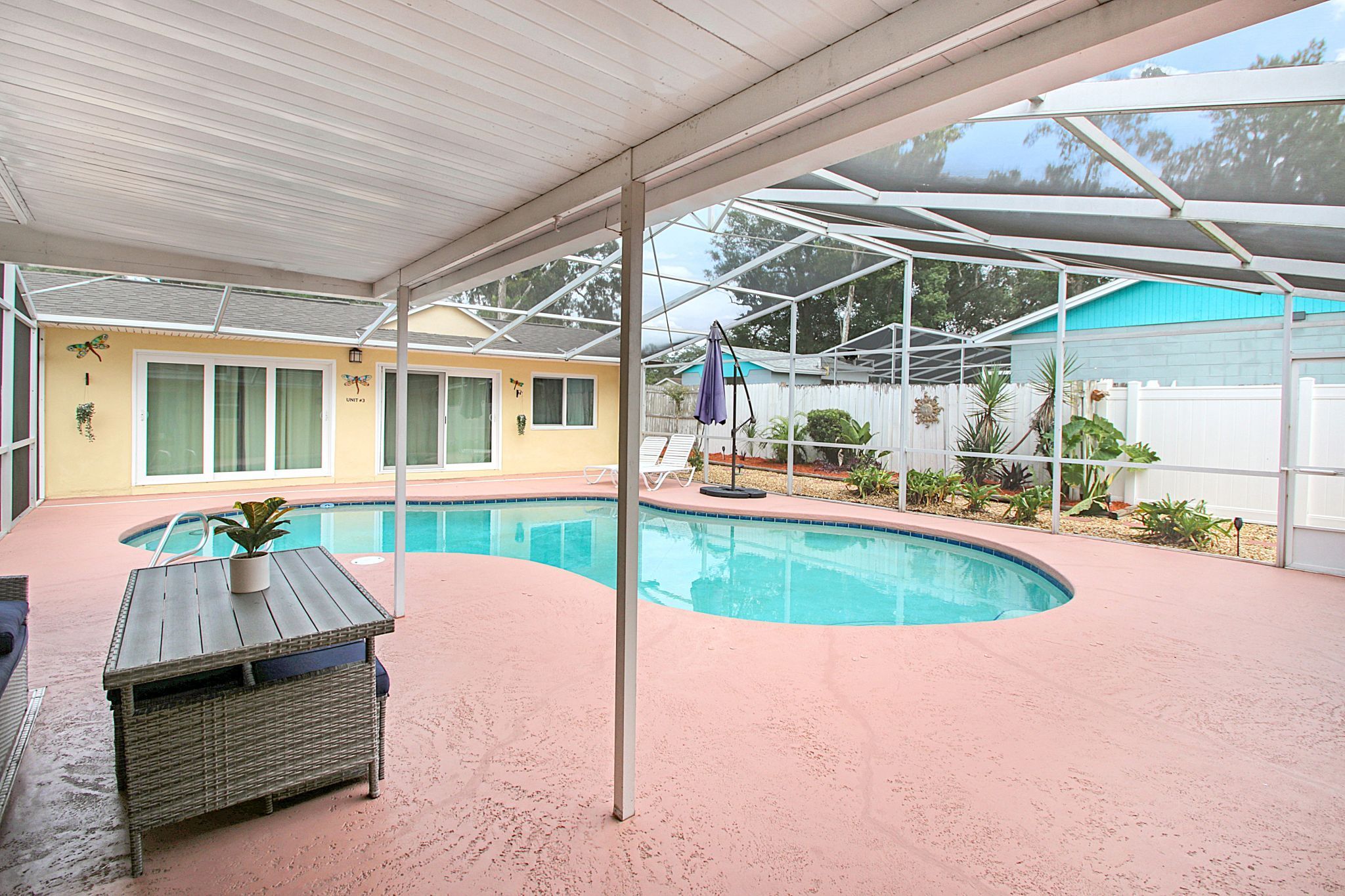 A covered outdoor swimming pool area with a lounge chair and a table, surrounded by a fence and vegetation.