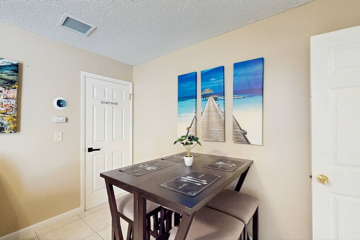 A dining area with a table and chairs, a door with a 'DO NOT ENTER' sign, and three beach-themed wall art pieces.