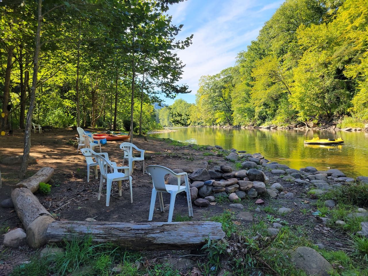Un espace extérieur paisible, aménagé avec des chaises en plastique et un banc en bois, entouré d’un paysage naturel composé d’arbres et d’une étendue d’eau calme.