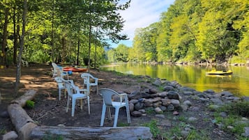 Un espace extérieur paisible, aménagé avec des chaises en plastique et un banc en bois, entouré d’un paysage naturel composé d’arbres et d’une étendue d’eau calme.