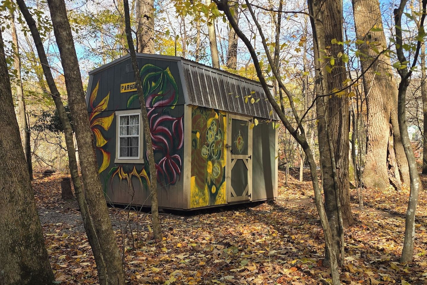 A small, rustic cabin with a colorful mural on its exterior, surrounded by trees and fallen leaves.