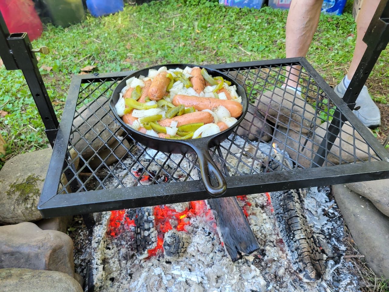 Una parrilla portátil con salchichas y verduras asándose sobre fuego abierto.
