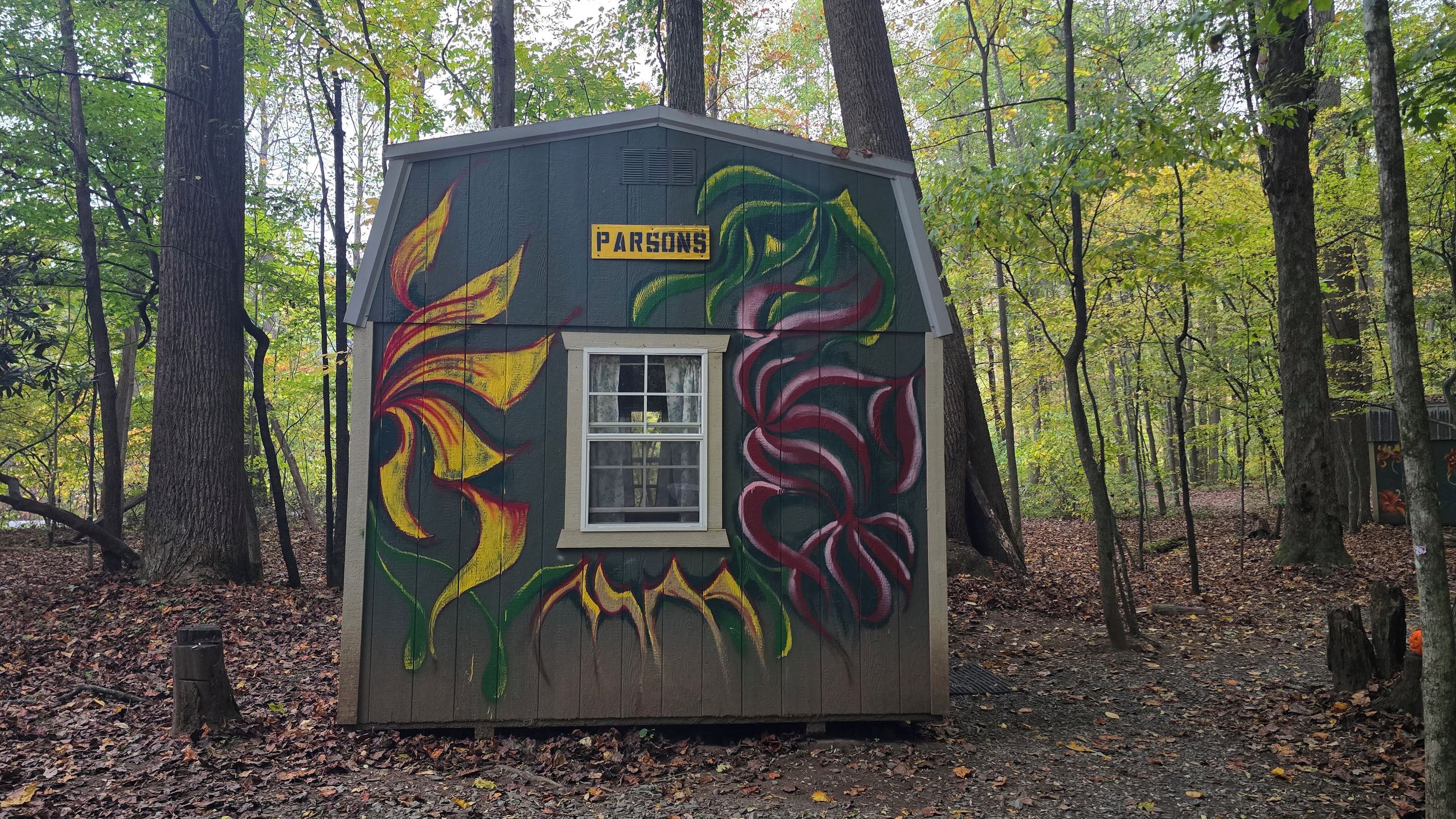 Una piccola baita di legno con una finestra e una porta, circondata da alberi e decorata con motivi a foglie colorate.