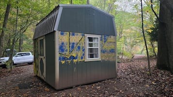 A small, single-story shed with a window, surrounded by trees and covered in graffiti.