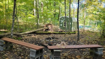 A forested area with a small cabin, two benches, and a fire pit.