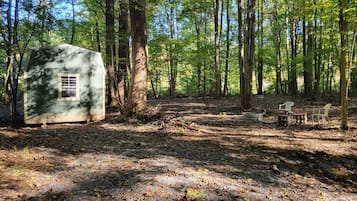 En enkel stuga i en skog med ett grusunderlag och en liten sittplats utomhus.
