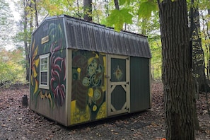 A small, rustic shed with a sloped roof, painted with abstract designs, situated in a wooded area.