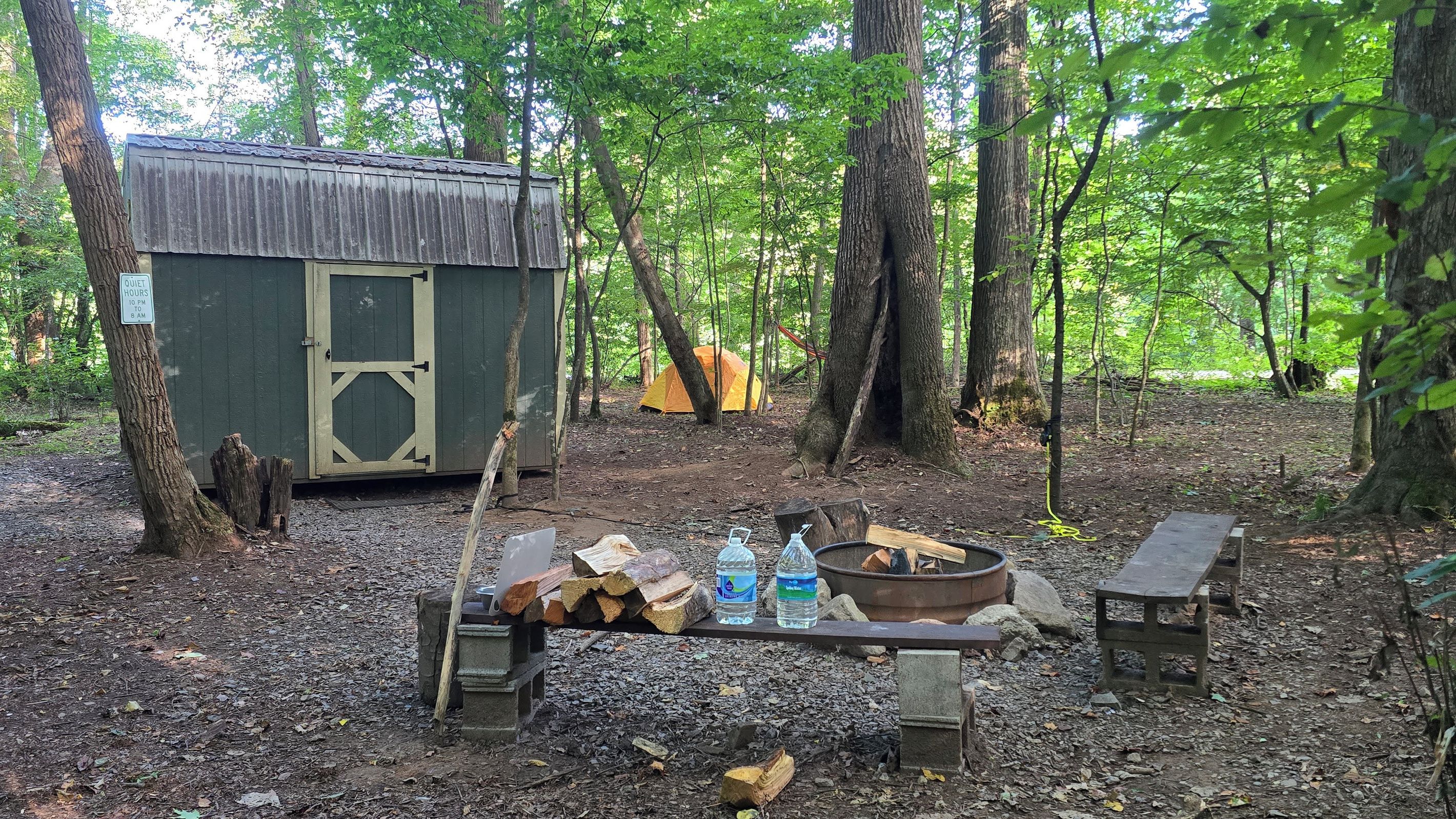 Ein rustikaler Campingplatz mit einer kleinen Holzhütte, einem Zelt und einer Feuerstelle, umgeben von Bäumen.
