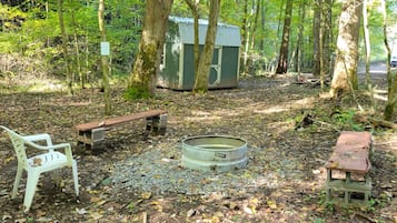 Eine ländliche Outdoor-Umgebung mit einem Feuerkorb aus Metall, einer Holzbank und einem kleinen Schuppen.