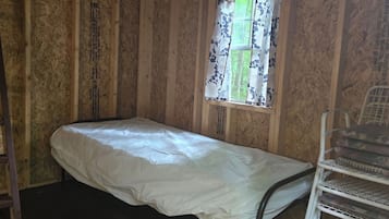A small, single-bed room with wooden walls, a window with floral curtains, and a metal chair.