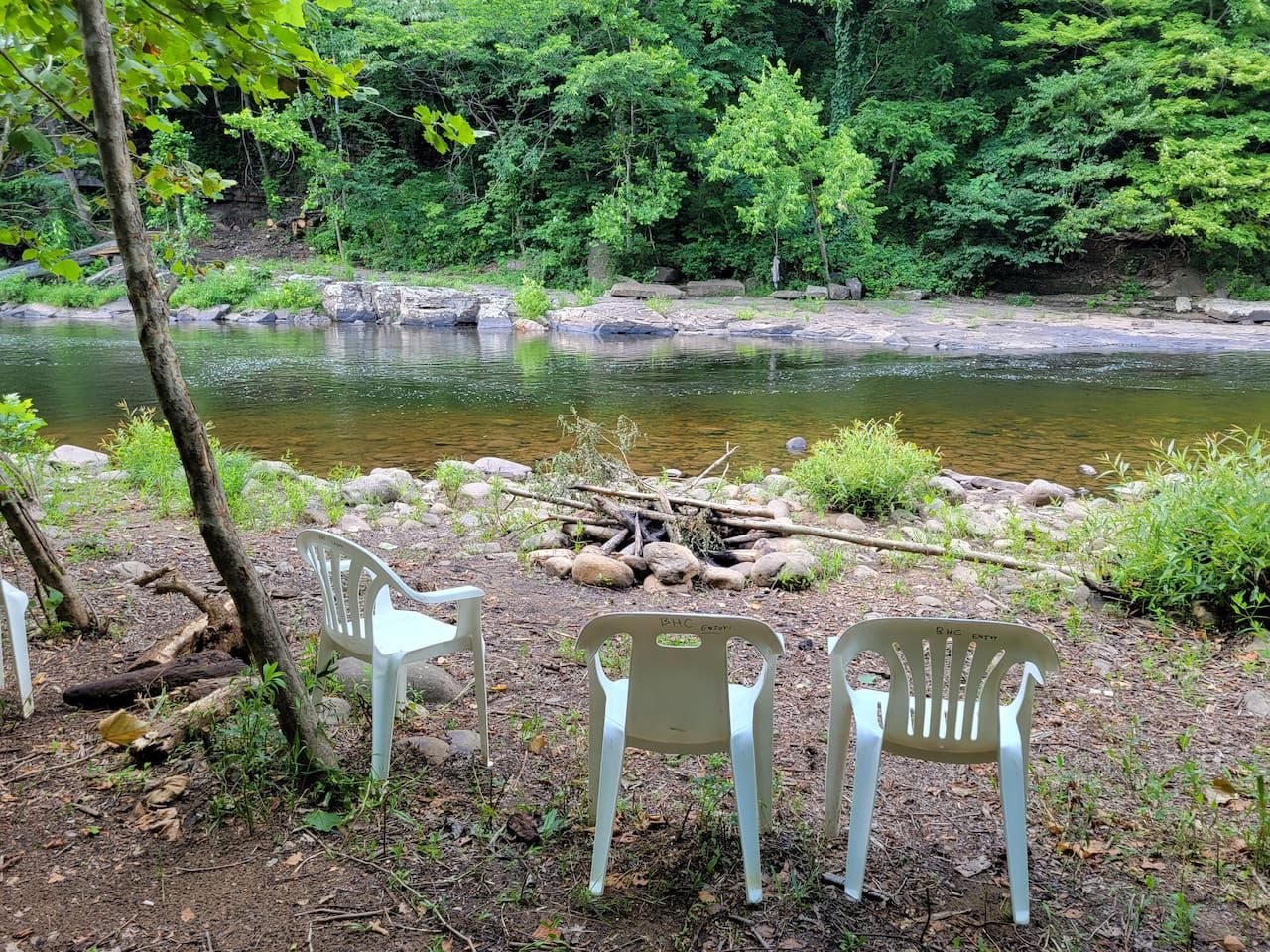 Eine natürliche Umgebung mit Fluss, Felsen und viel Grün. Drei weiße Plastikstühle stehen in der Nähe des Wassers.