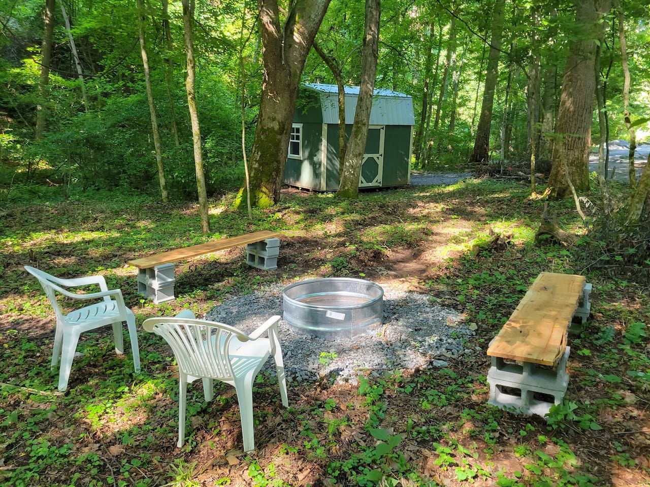 A campsite with a fire pit, two white plastic chairs, a wooden bench, and a small shed in a wooded area.