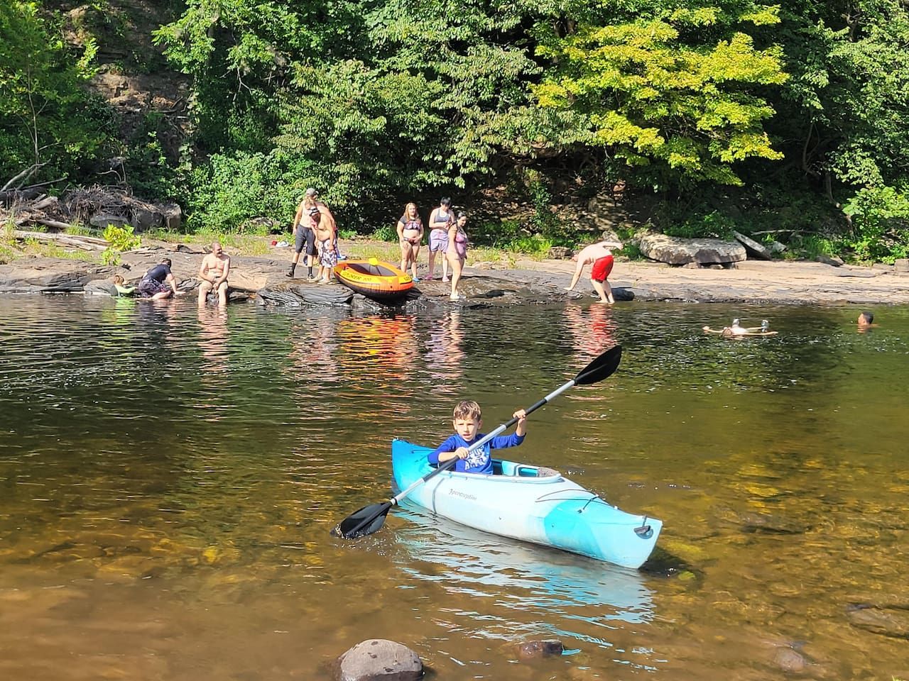 A kayaker in a blue kayak on a calm river with people on the shore.