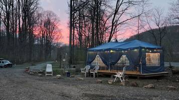 Ein blaues Zelt mit Tisch und Stühlen auf einer Schotterfläche, umgeben von Bäumen und einem Sonnenuntergang im Hintergrund.