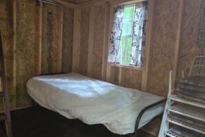 A small, single-bed room with wooden walls, a window with floral curtains, and a metal chair. - The Hendricks - Cabin CB3 (Parsons)