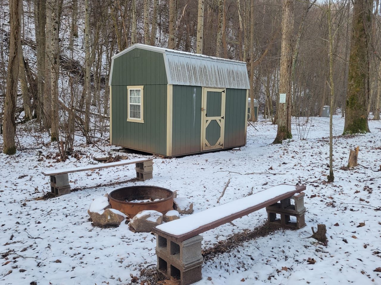 A wooden cabin with a fire pit and benches in a snowy forest.