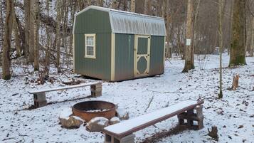 A wooden cabin with a fire pit and benches in a snowy forest.