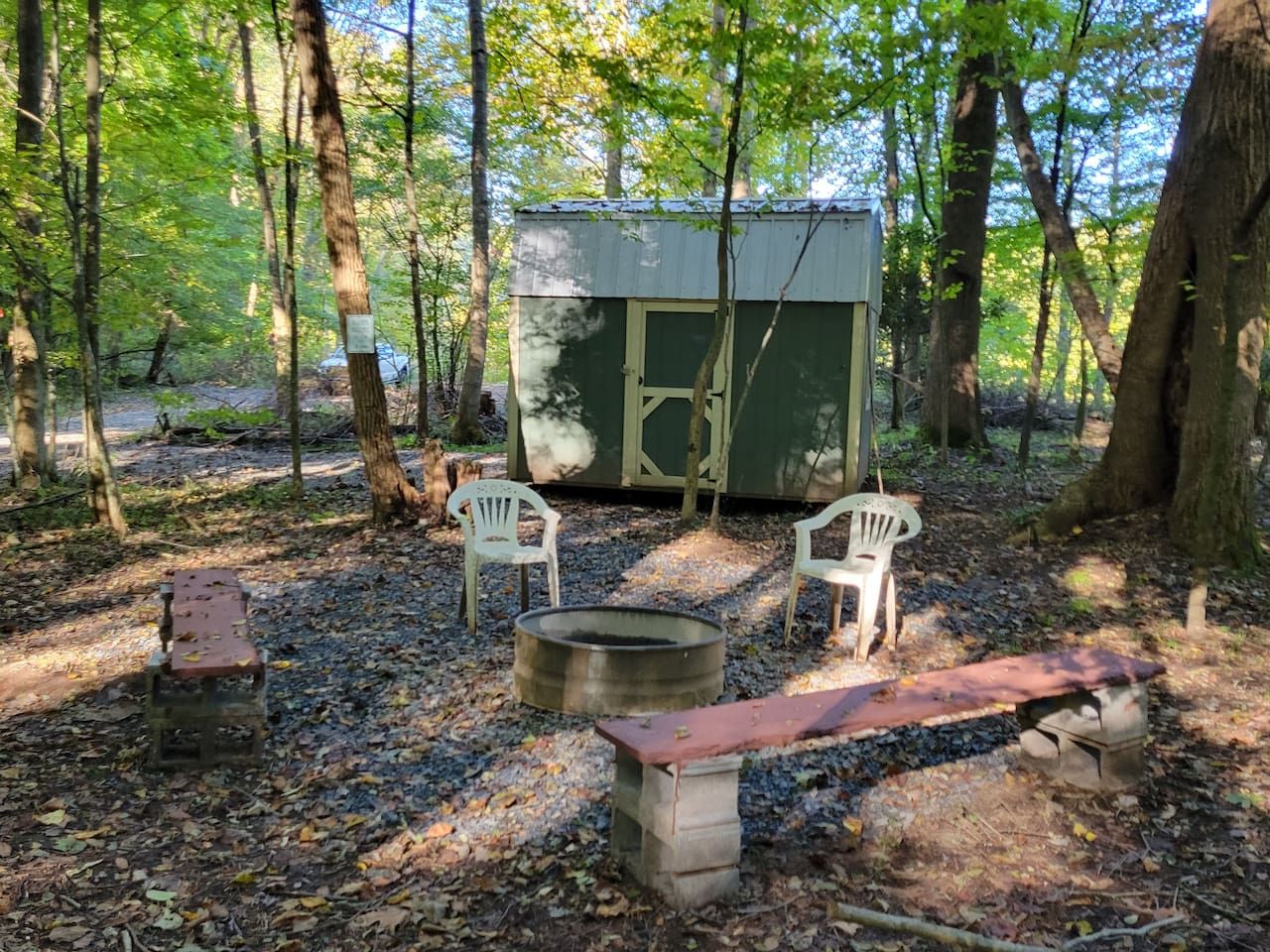 A rustic outdoor setting with a metal shed, two white plastic chairs, a fire pit, and a wooden bench.