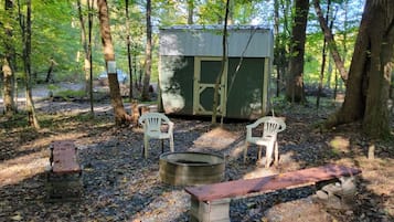 Een rustieke buitenomgeving met een metalen schuur, twee witte plastic stoelen, een vuurkorf en een houten bank.