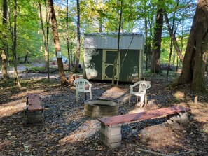 A rustic outdoor setting with a metal shed, two white plastic chairs, a fire pit, and a wooden bench. - The Hendricks - Cabin CB3 (Parsons)