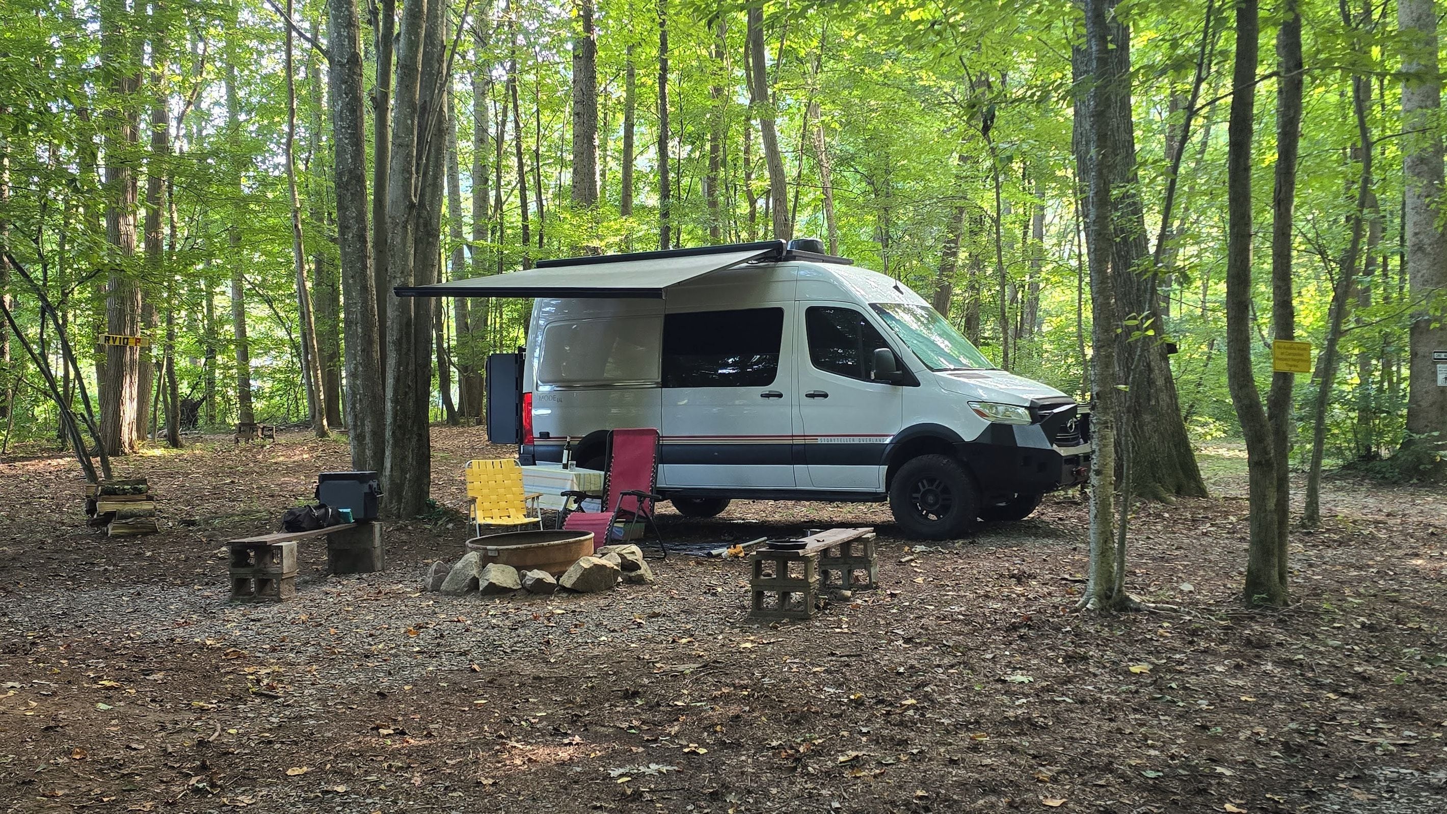 Una camioneta camper blanca estacionada en un bosque, con una mesa y sillas cerca.