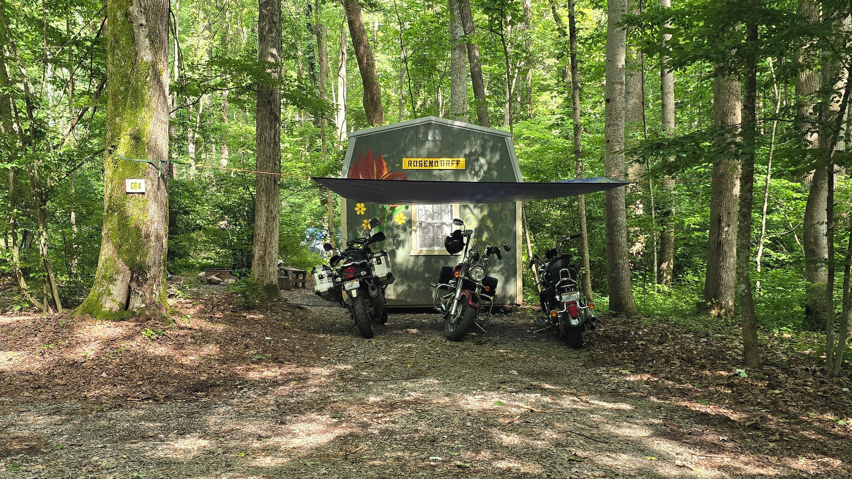 A small cabin with a sign reading 'ROSENDORFF' in a forested area with two motorcycles parked in front.