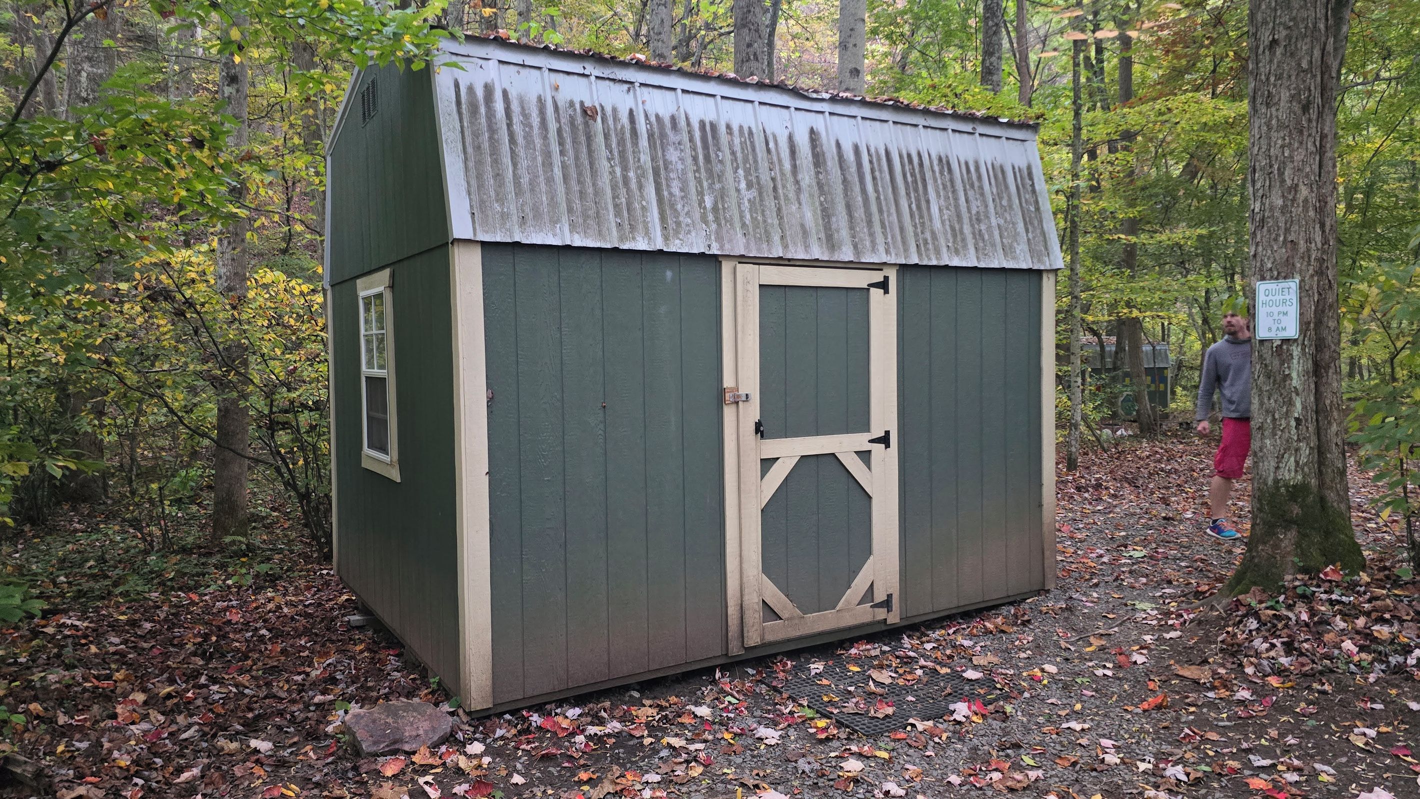 A small, single-story wooden shed with a metal roof, situated in a wooded area with fallen leaves on the ground.