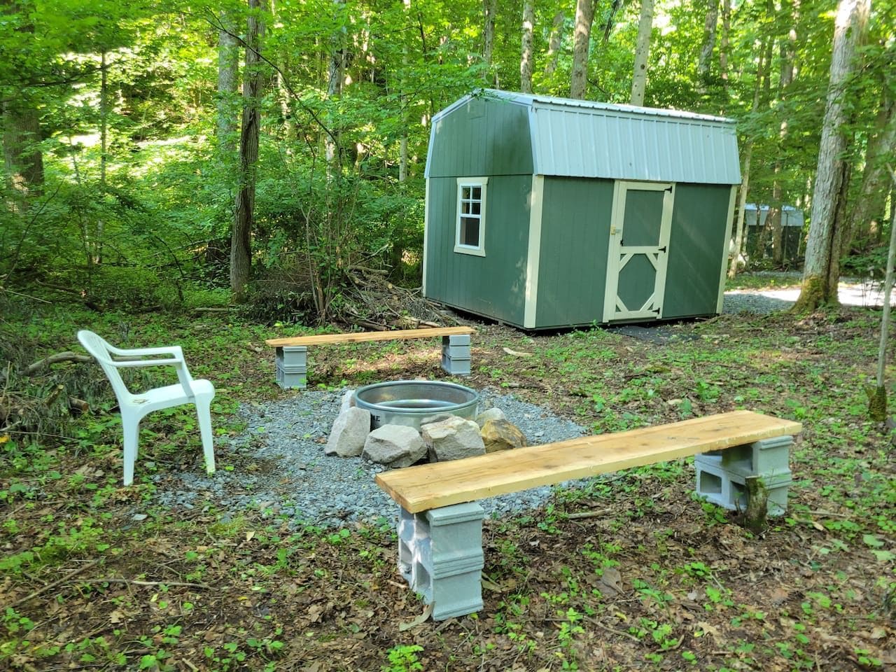 Un espace extérieur rustique aménagé avec une remise verte, une chaise en plastique blanche et deux bancs en bois.