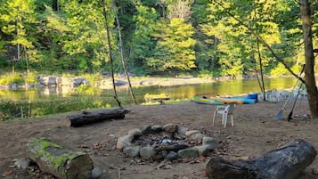 Ein Campingplatz mit Feuerstelle, Stühlen und Booten an einem von Bäumen gesäumten See.