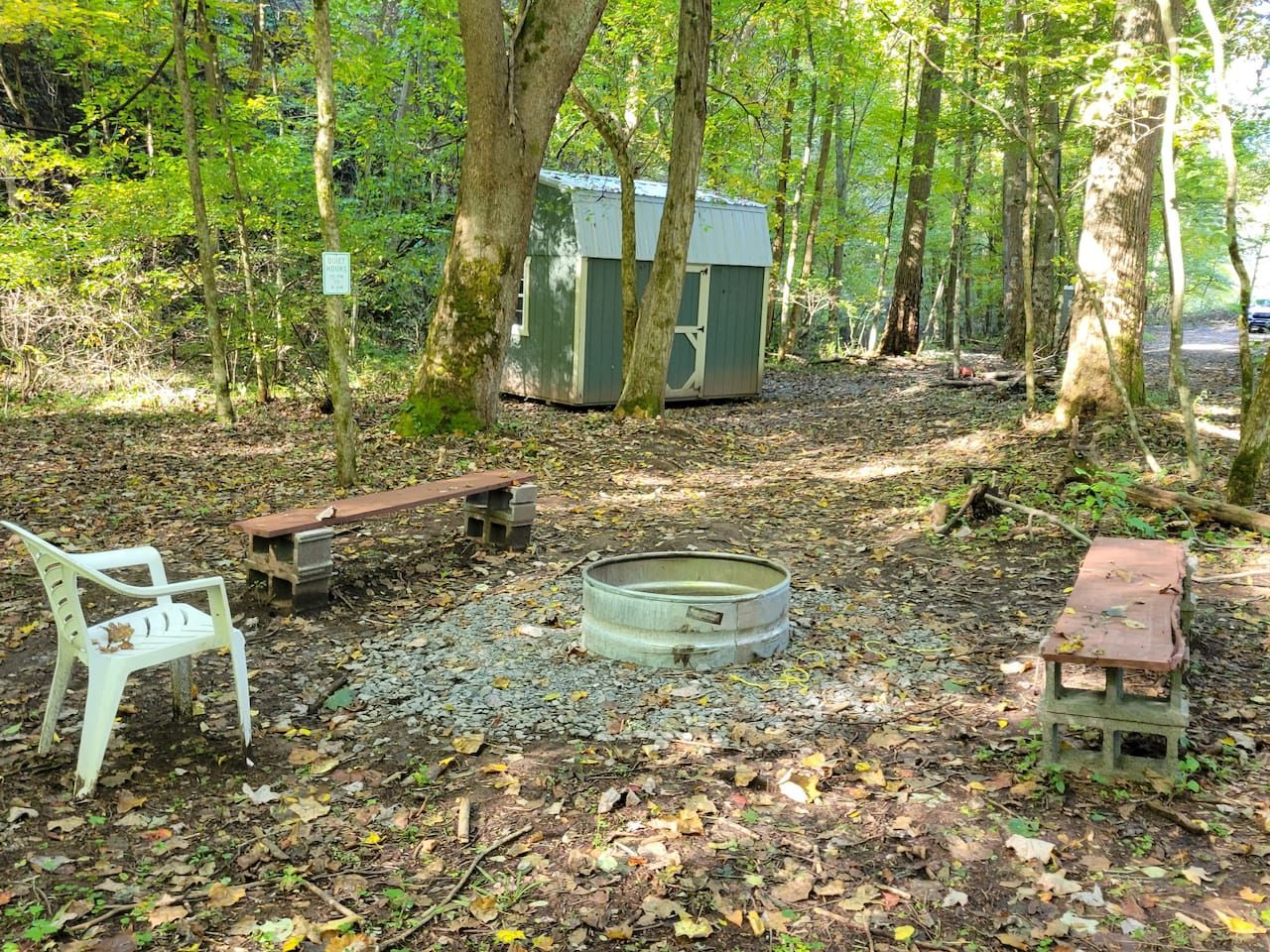 Eine ländliche Outdoor-Umgebung mit einem Feuerkorb aus Metall, einer Holzbank und einem kleinen Schuppen.