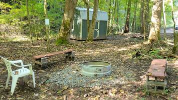 Eine ländliche Outdoor-Umgebung mit einem Feuerkorb aus Metall, einer Holzbank und einem kleinen Schuppen.