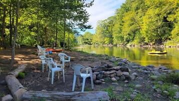 A serene outdoor seating area with plastic chairs and a log bench, surrounded by a natural landscape with trees and a calm body of water.