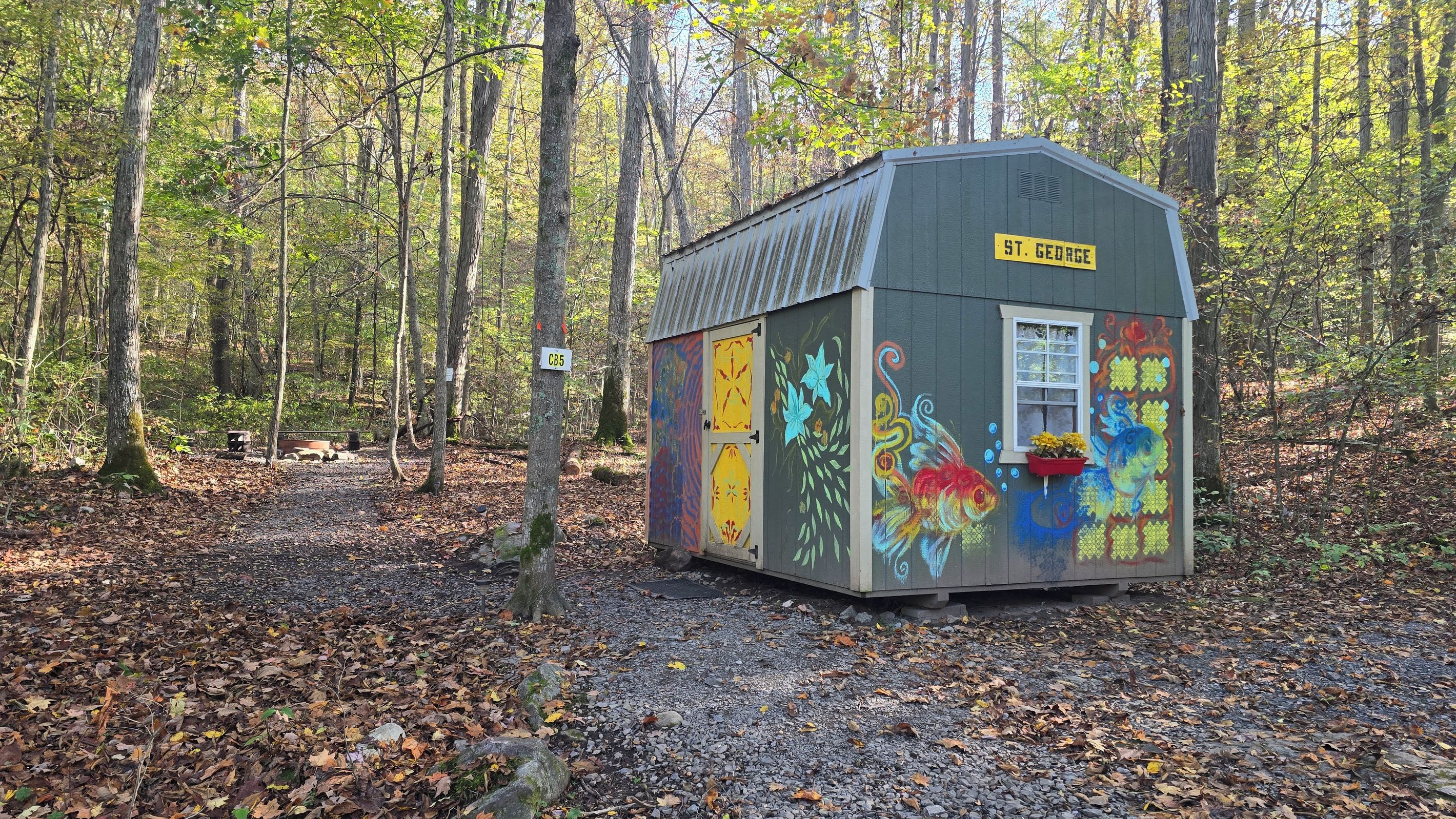 A small, colorful shed labeled 'St. George' in a forested area with a gravel path.
