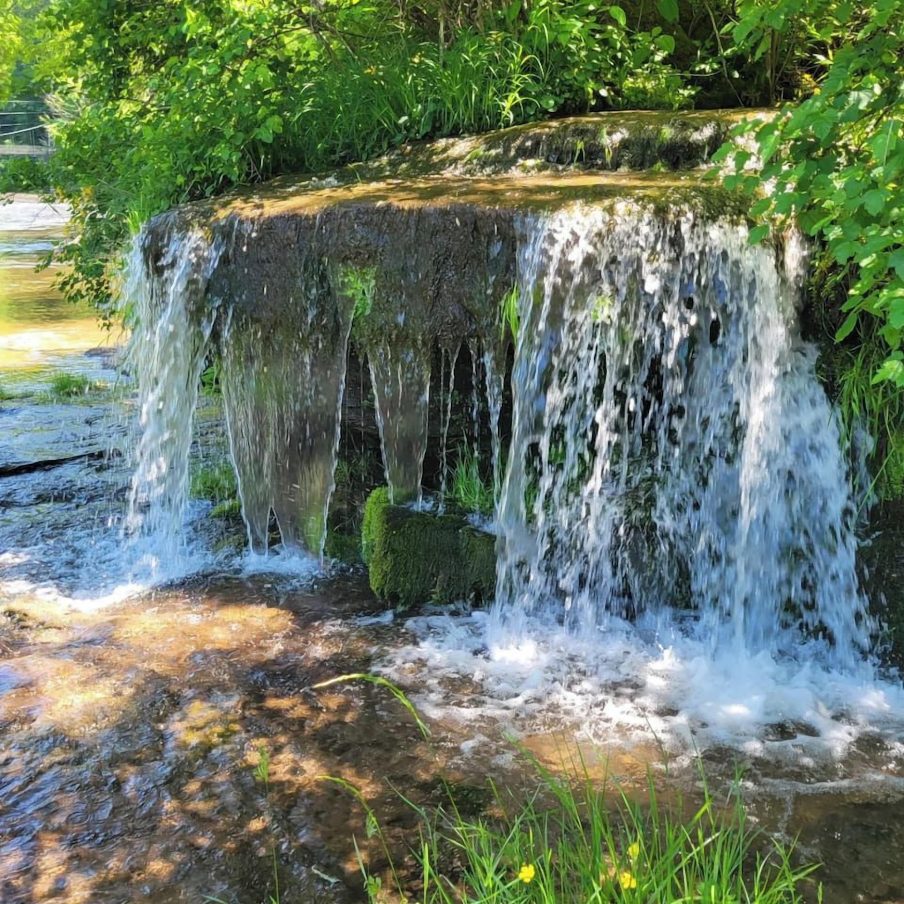 A waterfall cascading over a moss-covered rock structure surrounded by lush greenery.