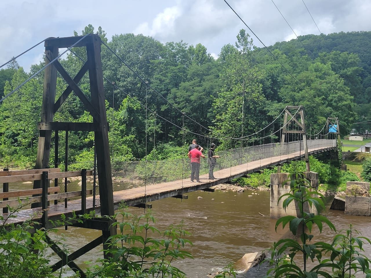 Eine Hängebrücke über einem Fluss mit Metallfachwerk und Holzdielen, umgeben von viel Grün.