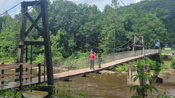 Eine Hängebrücke über einem Fluss mit Metallfachwerk und Holzdielen, umgeben von viel Grün.