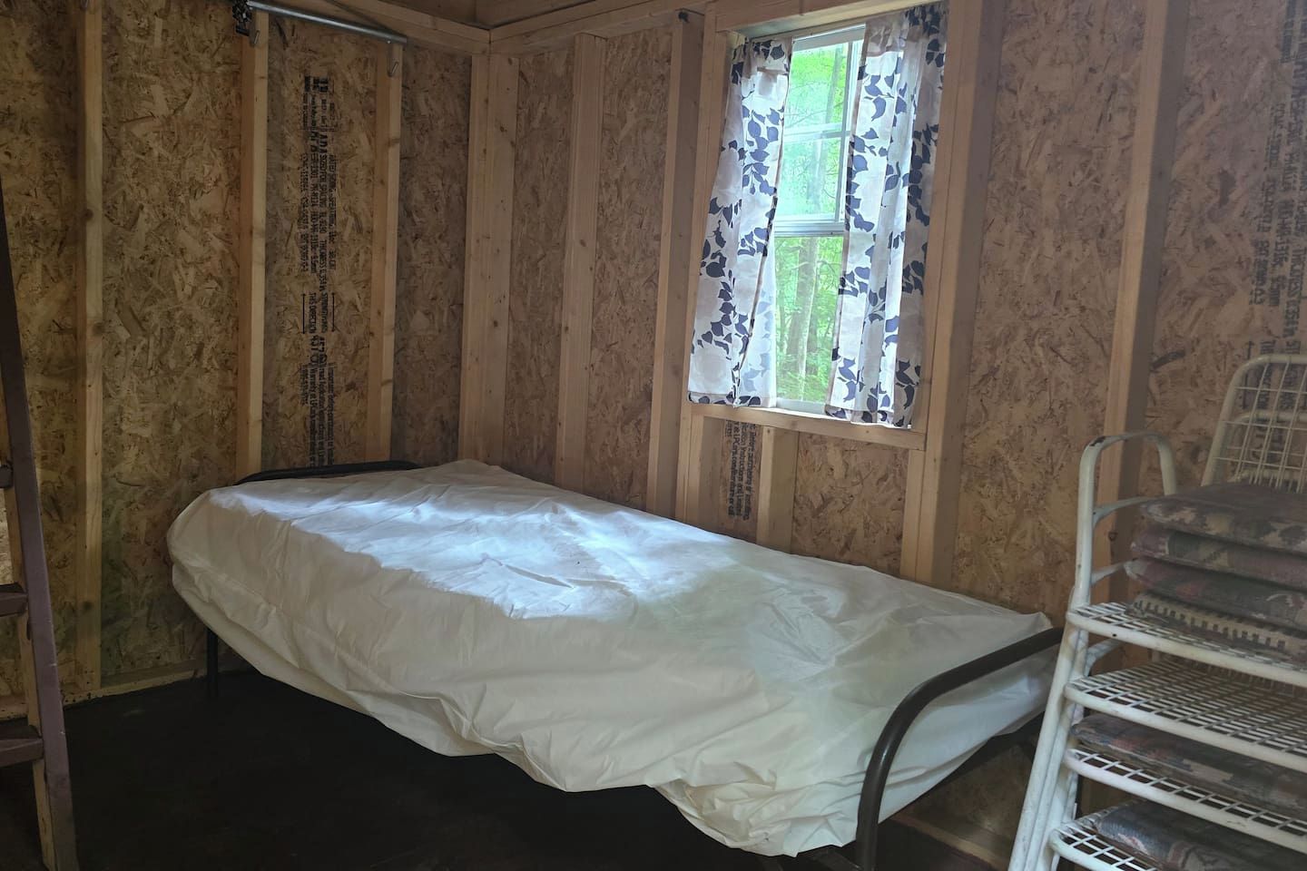 A small, single-bed room with wooden walls, a window with floral curtains, and a metal chair.