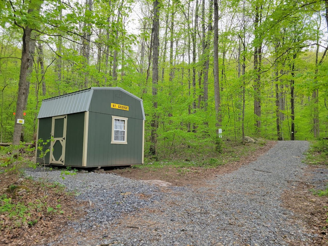 A small, single-story building with a door and window, surrounded by a forested area with a gravel path leading to it.