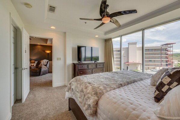 A bedroom with a large bed, a ceiling fan, a TV, and a view of a building outside.