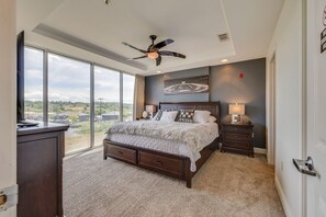 A bedroom with a large bed, a ceiling fan, a nightstand, and a view of the outdoors through large windows.