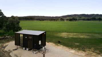 A small, dark-colored, single-story structure with a flat roof, possibly a shed or storage unit, situated on a gravel surface with a grassy field and hills in the background.