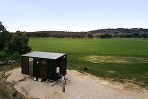 Uma construção pequena e escura, de um andar, com telhado plano, possivelmente um depósito ou arrecadação, localizada em terreno de cascalho, com um campo gramado e colinas ao fundo.