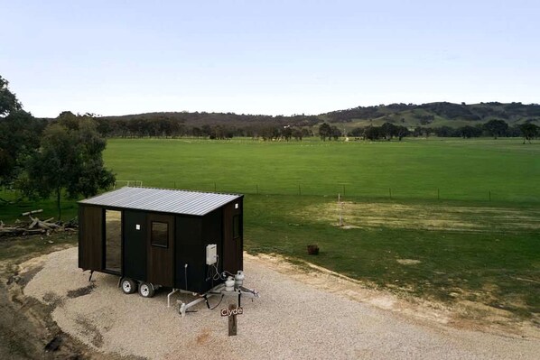 A small, dark-colored, single-story structure with a flat roof, possibly a shed or storage unit, situated on a gravel surface with a grassy field and hills in the background.