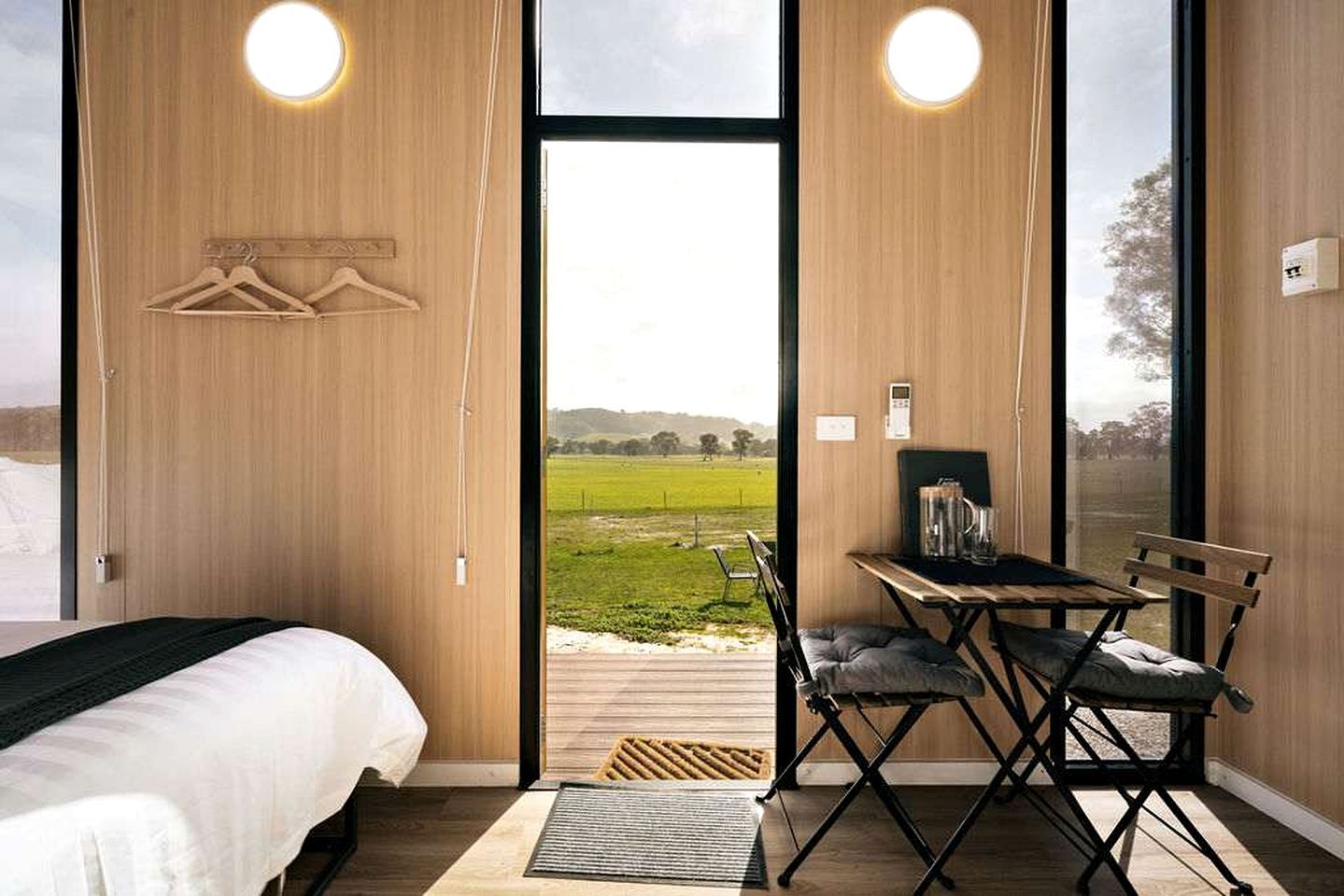 Ein Schlafzimmer mit einem Bett, einem Schreibtisch mit Stuhl und Blick auf ein grünes Feld durch ein großes Fenster.