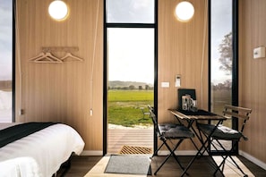 A bedroom with a bed, a desk with a chair, and a view of a green field through a large window.