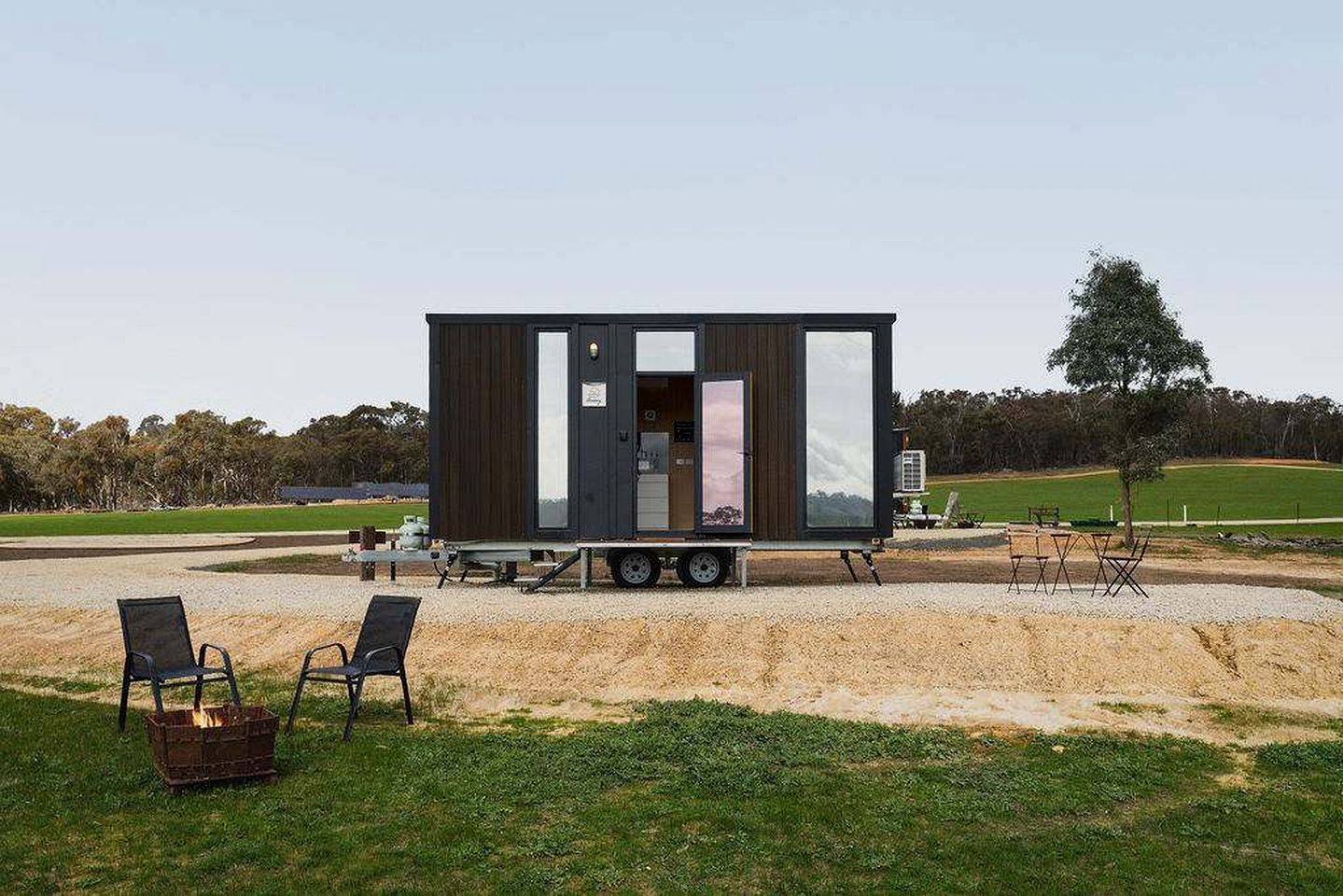 Una casa móvil moderna y compacta con un amplio ventanal corredera, ubicada en una zona de grava con mobiliario de exterior.