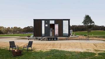 Una casa móvil moderna y compacta con un amplio ventanal corredera, ubicada en una zona de grava con mobiliario de exterior.