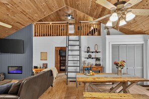 Ein Wohnraum im Loft-Stil mit Holzbalkendecke, Schlafplatz in der Mansarde, Esstisch und Fernseher.