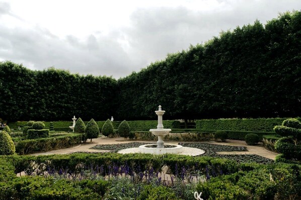 A formal garden with a central fountain, trimmed hedges, and a variety of plants.